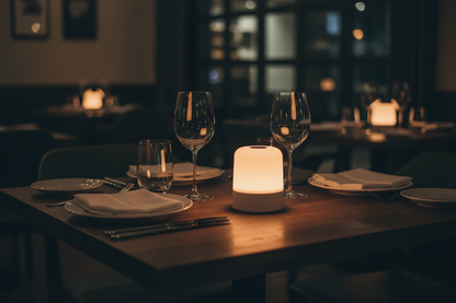 Small round white portable rechargeable table lamp glowing warm amber on restaurant dining table set for two in dark moody atmosphere